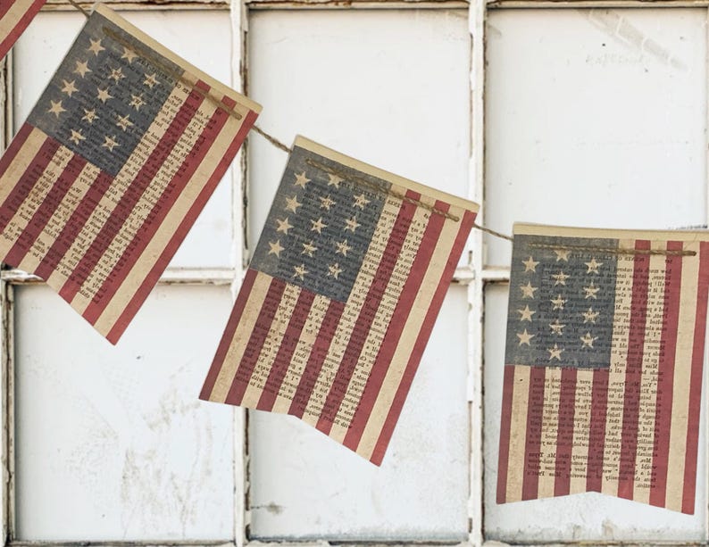 May include: A decorative bunting banner featuring rectangular flags printed with the American flag design. Each flag has a blue canton with white stars and red and white stripes, with the entire surface overlaid with vintage-style printed text.