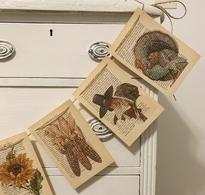 May include: Thanksgiving-themed garland featuring vintage book pages with images of a turkey, corn, and a pilgrim. The garland is strung on twine and hangs against a white dresser.