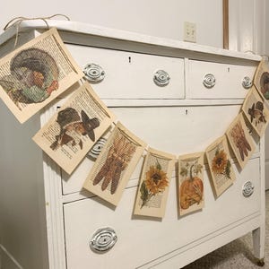 May include: Thanksgiving-themed banner featuring vintage book pages with turkey, pilgrim, corn, sunflower, and pumpkin illustrations. The banner is strung on twine and hangs in front of a white dresser. A festive decoration for the fall season.