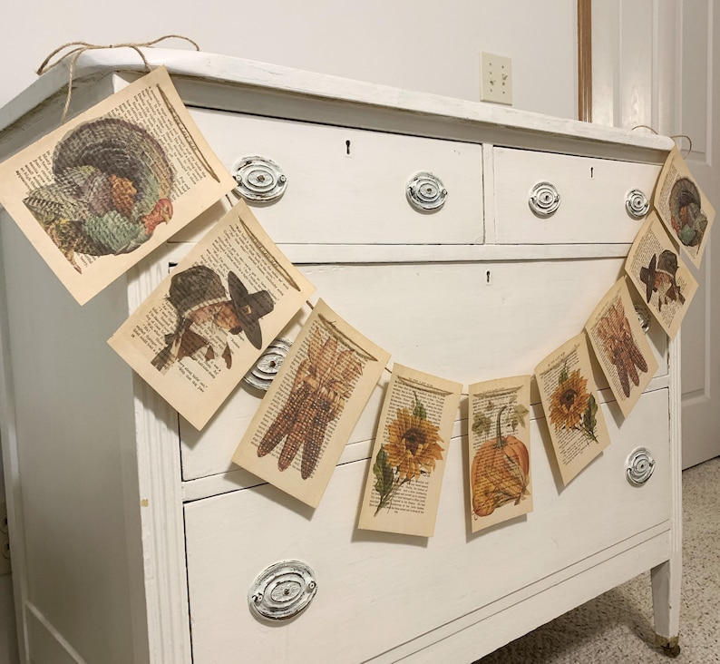 May include: Thanksgiving-themed banner featuring vintage book pages with turkey, pilgrim, corn, sunflower, and pumpkin illustrations. The banner is strung on twine and hangs in front of a white dresser. A festive decoration for the fall season.