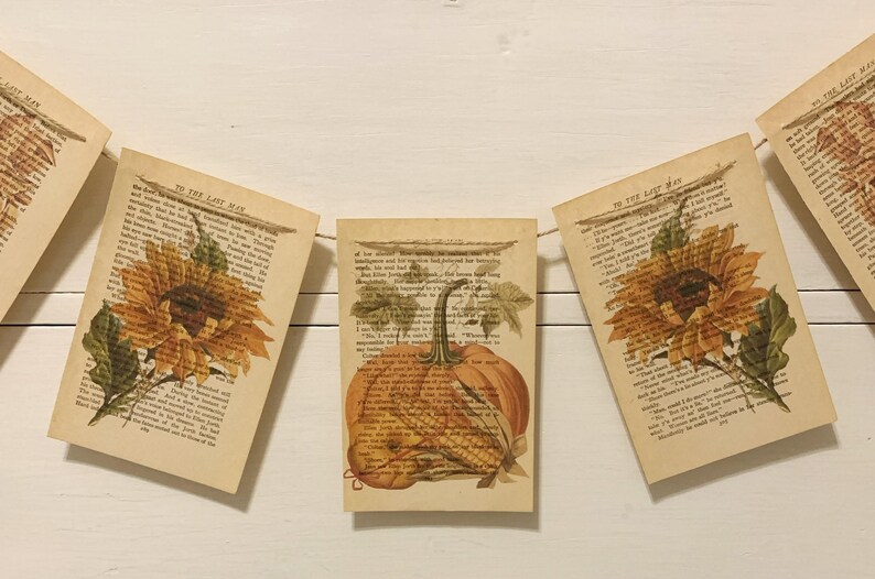 May include: A garland of vintage book pages with autumn-themed illustrations. The banner features images of sunflowers and a pumpkin, printed on aged paper. The garland is strung on a thin twine, perfect for fall decor.