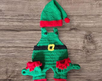 May include: A green and red crochet elf outfit, comprising a hat and dungarees. The hat has a red brim and a red pom-pom. The dungarees feature a black belt with a yellow buckle and red trim on the legs. The outfit is designed for a baby.