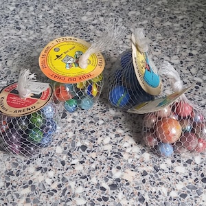 May include: Four mesh bags filled with glass marbles in various colours and sizes. Each bag features a circular cardboard header tag with printed text including brand names like Arend, Bon Jour, and Corsaire, and size labels such as 16 mm and 25 mm.
