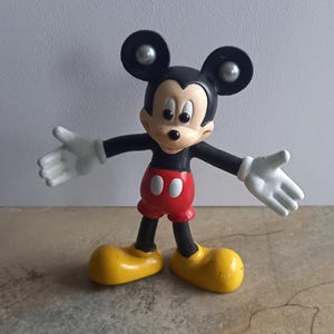 Könnte beinhalten: Eine schwarze und rote Mickey Mouse Figur aus Plastik mit großen, runden Ohren und einem breiten Lächeln. Die Figur hat silberne Knöpfe an den Ohren und trägt gelbe Schuhe.