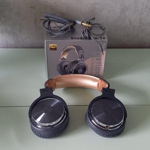 May include: Studio Pro 10 DJ headphones with a tan padded headband and dark gray ear cups featuring metallic circular accents. The headphones are positioned in front of their original gray packaging box, which includes a coiled audio cable with a gold plug.