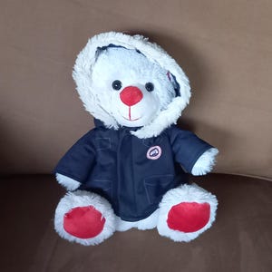 Op de afbeelding: Een witte pluche teddybeer met een marineblauw jack met capuchon en imitatiebont. De beer heeft een felrode neus en rode stoffen zolen onder de voeten. Op de borst van het jack is een klein, rond logopatch zichtbaar.