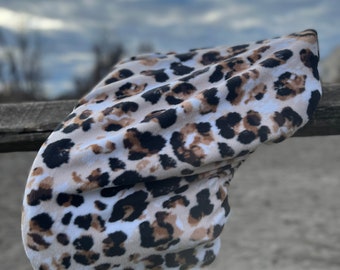 May include: A soft, leopard print headband in shades of brown, black, and cream. The fabric is draped over a wooden fence, with a blurred background of a cloudy sky.
