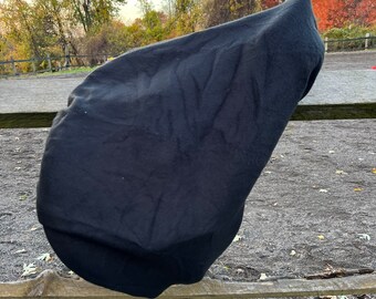 May include: Black saddle cover hanging on a wooden fence. The cover is made of a soft, textured fabric. The background is a blurry image of trees and a field.