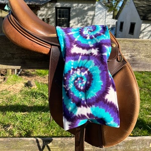 May include: A purple and turquoise tie-dye fleece saddle pad hanging on a wooden fence.