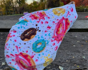 May include: A white headband with a colorful donut pattern. The donuts are decorated with pink, brown, blue, and yellow frosting and sprinkles. The headband is made of a soft, stretchy material.