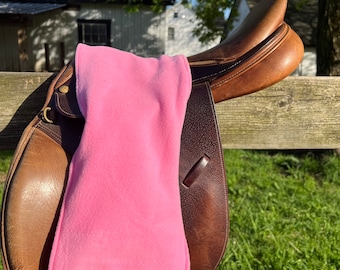 May include: A pink fleece saddle pad for a horse saddle. The saddle pad is on a brown leather saddle.