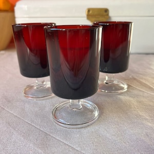 Peut inclure: Trois petits verres à shot en verre transparent avec une base rouge foncé. Les verres sont disposés en rangée sur une nappe en lin blanc.