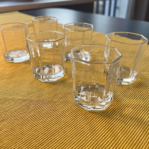 May include: Six clear glass shot glasses on a yellow striped surface. The glasses are hexagonal in shape and have a rounded bottom.