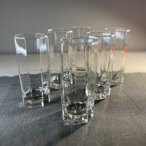 Peut inclure: Six verres à boire en verre transparent avec un design géométrique facetté. Les verres sont disposés en demi-cercle sur une surface texturée grise.