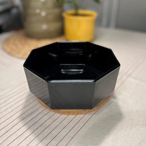 Peut inclure: Un bol noir octogonal avec une base en liège. Le bol a une finition brillante et repose sur un dessous de verre rond en liège. L'arrière-plan comprend une table avec un motif à rayures rouges et blanches et quelques plantes en pot.