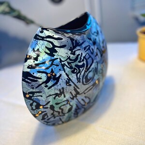Peut inclure: Un vase en céramique en forme de croissant arrondi avec une finition multicolore dans des tons de bleu, vert et argent. La surface est ornée d'un motif craquelé noir et d'éclaboussures noires abstraites, accentuées par de petits points dorés.
