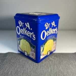 Könnte beinhalten: Eine blaue Blechdose mit dem Text "Dr. A. Oetker's Pudding" darauf. Die Dose hat ein Bild von einem gelben Pudding darauf.