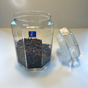 Peut inclure: Un bocal en verre clair, octogonal, rempli de grains de café brun foncé. Le couvercle est sur le côté. Le bocal porte un logo Luminarc bleu et jaune sur le devant.