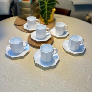 Peut inclure: Un ensemble de six tasses à thé et soucoupes en céramique blanche. Les tasses présentent un design géométrique à facettes et reposent sur des soucoupes octogonales assorties. L'ensemble est disposé sur une nappe de couleur claire.