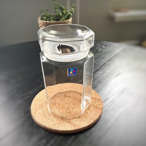 May include: A clear, octagonal glass jar with a matching lid, on a round cork coaster. The jar has a white rim and a blue and yellow logo. A small plant in a woven basket is in the background.