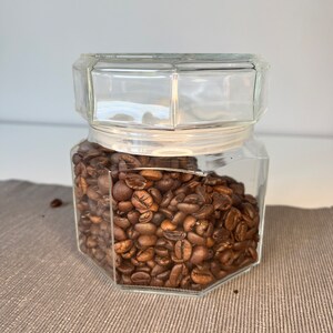 Peut inclure: Un bocal en verre transparent de forme hexagonale rempli de grains de café torréfiés. Le bocal a un couvercle et est posé sur une surface texturée brune.