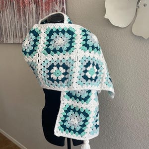 May include: A white crocheted scarf with a green, blue, and white granny square pattern. The scarf has a white tassel.