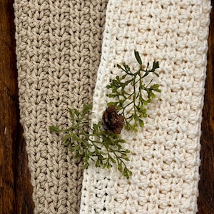May include: Close-up of two crocheted cloths, one beige and one cream, with a sprig of greenery and a small pinecone. The cloths have a textured, handmade appearance. The image highlights the natural, rustic aesthetic of the textiles.