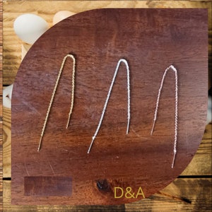 Può includere: Tre paia di orecchini a filo in oro, argento e oro rosa. Ogni orecchino ha una catena sottile che pende dall'orecchio. Gli orecchini sono esposti su una superficie di legno scuro. Il testo "D&A" è visibile.