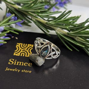 May include: A silver ring with a Celtic knot design and an abalone shell inlay. The ring is on a black card with the text "Simee Jewelry store".
