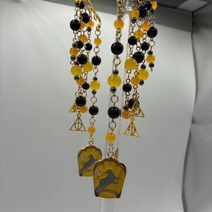 Puede incluir: Un par de pendientes tipo ear cuff dorados con cadenas en cascada adornadas con cuentas amarillas y negras. Cada pendiente incluye pequeños dijes triangulares dorados con un símbolo circular y un colgante amarillo en forma de escudo.