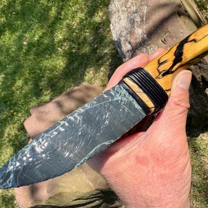 May include: A handcrafted knife with a gray, patterned blade and a light-colored wooden handle. The handle features black accents and is bound with black material. The knife is held in a hand against a backdrop of green grass and a tree trunk.