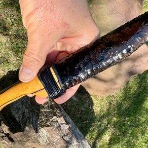 May include: A handcrafted obsidian knife with a light wood handle. The blade is a deep, glossy black, and the handle is wrapped with black cord. The knife is held against a backdrop of green grass and a weathered log.