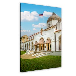 May include: A photograph of the historic Quapaw Baths building, featuring a white facade with arched entryways and a domed roof. The American flag flies atop the building under a blue sky with fluffy clouds. The words "QUAPAW BATHS" are visible.