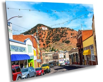 May include: A street scene in a small town with colorful buildings and a red rock hill in the background. The street is lined with parked cars and there are string lights strung across the street. The sign on the hill says "Welcome to Silver City".
