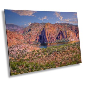 May include: A scenic landscape photograph featuring a large body of water surrounded by red rock cliffs and desert vegetation. The water is a deep blue and the sky is a bright blue with white clouds. The cliffs are a reddish-brown color and are covered in green vegetation.