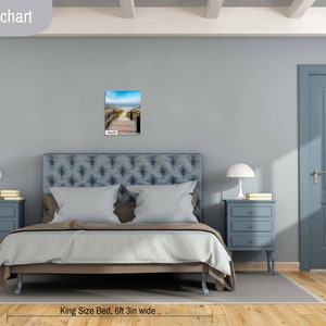 May include: A bedroom with a king-size bed, two bedside tables with lamps, and a door. The bed has a blue headboard and a white duvet. The walls are light blue. A framed photograph of a beach scene hangs on the wall above the headboard. The caption below the bed reads "King Size Bed, 6ft 3in wide".