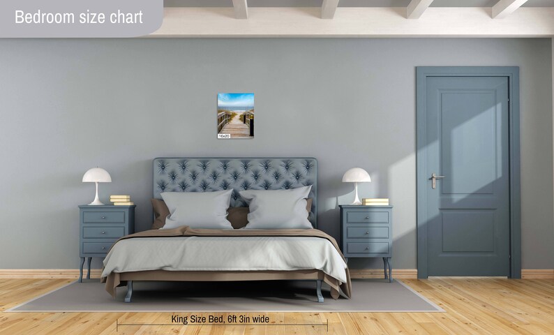 May include: A bedroom with a king-size bed, two bedside tables with lamps, and a door. The bed has a blue headboard and a white duvet. The walls are light blue. A framed photograph of a beach scene hangs on the wall above the headboard. The caption below the bed reads "King Size Bed, 6ft 3in wide".
