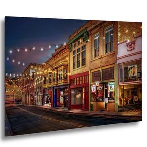 May include: A vibrant street scene at dusk, featuring a row of historic buildings with colorful storefronts. String lights illuminate the street, and the sky transitions from blue to dark. The buildings are made of brick and have various architectural details.