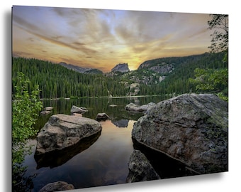 May include: A scenic landscape photograph of a lake surrounded by mountains and evergreen trees. The sky displays a colorful sunset with shades of orange and yellow. Large rocks are in the foreground, and the water reflects the sky and surrounding scenery.