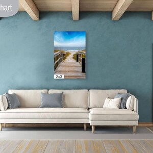 May include: A white sectional sofa with cushions in a living room with a blue wall and a wooden door. A framed photograph of a wooden walkway leading to the beach hangs on the wall. The photograph is labelled "2x3".