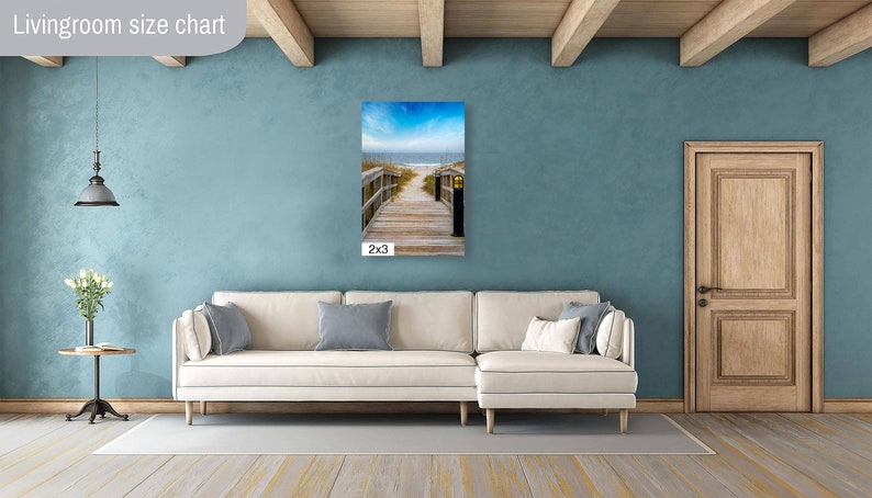 May include: A white sectional sofa with cushions in a living room with a blue wall and a wooden door. A framed photograph of a wooden walkway leading to the beach hangs on the wall. The photograph is labelled "2x3".