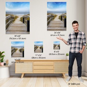 May include: Four different sizes of canvas prints featuring a wooden walkway leading to a beach with a blue sky and ocean in the background. The prints are shown in a room with a wooden table and two people standing next to the prints.