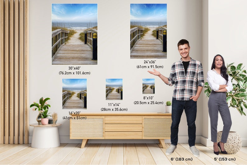 May include: Four different sizes of canvas prints featuring a wooden walkway leading to a beach with a blue sky and ocean in the background. The prints are shown in a room with a wooden table and two people standing next to the prints.