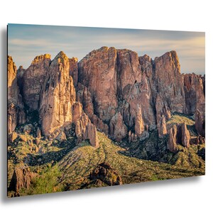 May include: A landscape photograph of a mountain range with rugged, reddish-brown rock formations. The mountains are illuminated by sunlight, with shadows creating depth. Green vegetation covers the lower slopes, and a blue sky is visible.