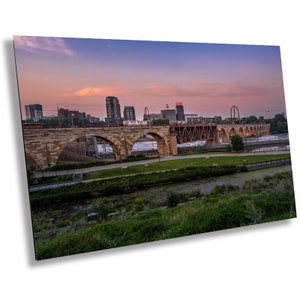 May include: A stone arch bridge over a river with a city skyline in the background. The sky is a vibrant pink and purple at sunset.