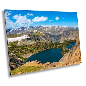 May include: A panoramic view of a mountain lake in a rugged landscape. The lake is a deep blue, surrounded by green hills and snow-capped mountains. The sky is a bright blue with fluffy white clouds.