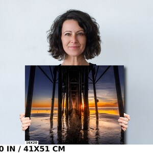 May include: A photograph of a sunset over the ocean with a pier in the foreground. The pier is made of wood and has a long walkway that leads out to the water. The sky is a mix of orange, pink, and purple, and the water is a deep blue. The image is 16x20 inches or 41x51 centimeters.