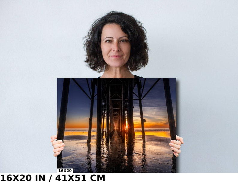 May include: A photograph of a sunset over the ocean with a pier in the foreground. The pier is made of wood and has a long walkway that leads out to the water. The sky is a mix of orange, pink, and purple, and the water is a deep blue. The image is 16x20 inches or 41x51 centimeters.