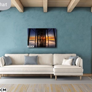 May include: A living room with a light blue wall, a white couch, and a wooden door. A photograph of a sunset under a pier is hanging on the wall. The image is labeled "24x36 in / 61x91 cm".