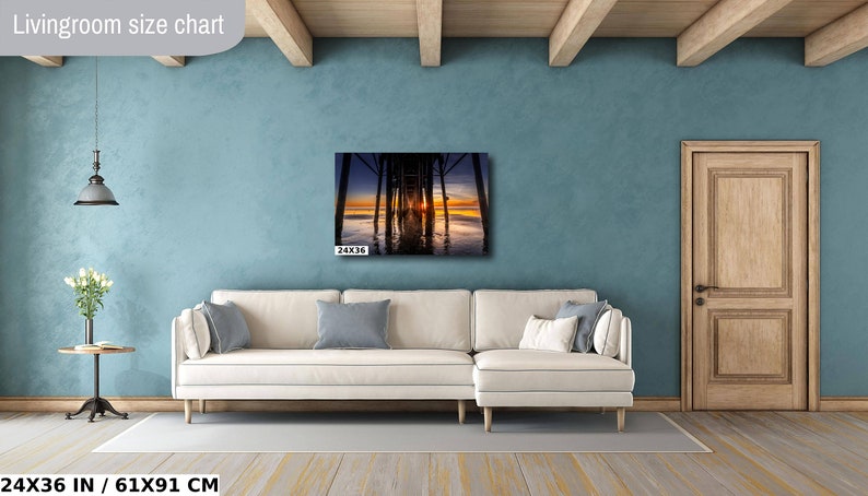 May include: A living room with a light blue wall, a white couch, and a wooden door. A photograph of a sunset under a pier is hanging on the wall. The image is labeled "24x36 in / 61x91 cm".